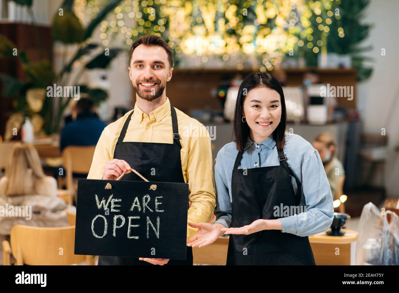 Willkommen, wir sind OFFEN. Zwei multirassische freundliche Kellner in Uniform stehen in einem Restaurant, Café oder Bar, zeigen das Schild OFFEN, mit einem angenehmen Lächeln. Teamwork-Konzept Stockfoto