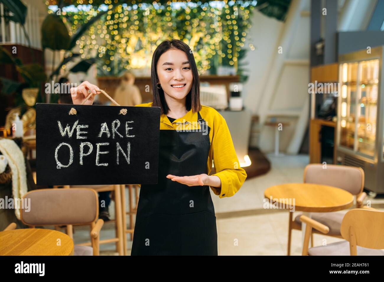 Wir sind offen. Portrait einer glücklichen hübschen asiatischen Kellnerin, die drinnen in einem Restaurant oder Café mit schwarzer Schürze steht, hält EIN Schild OFFEN und lächelt freundlich Stockfoto