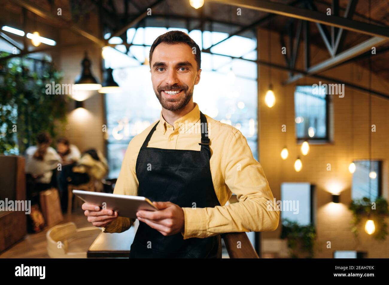 Attraktive glücklich kaukasischen männlichen Kellner oder Barista trägt schwarze Schürze steht in einem Restaurant, Café oder Bar, hält ein Menü und mit freundlichem Lächeln schaut auf die Kamera Stockfoto