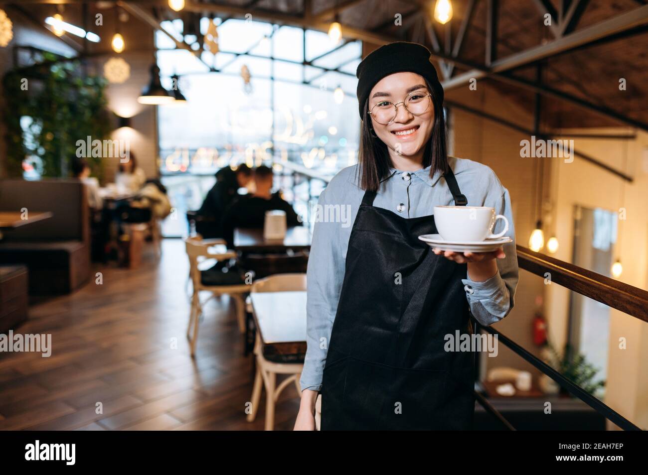 Schöne glückliche asiatische Kellnerin oder Barista mit Brille steht in einem Restaurant, Café oder Bar, hält eine Tasse aromatischen Kaffee und mit freundlichem Lächeln schaut auf die Kamera Stockfoto