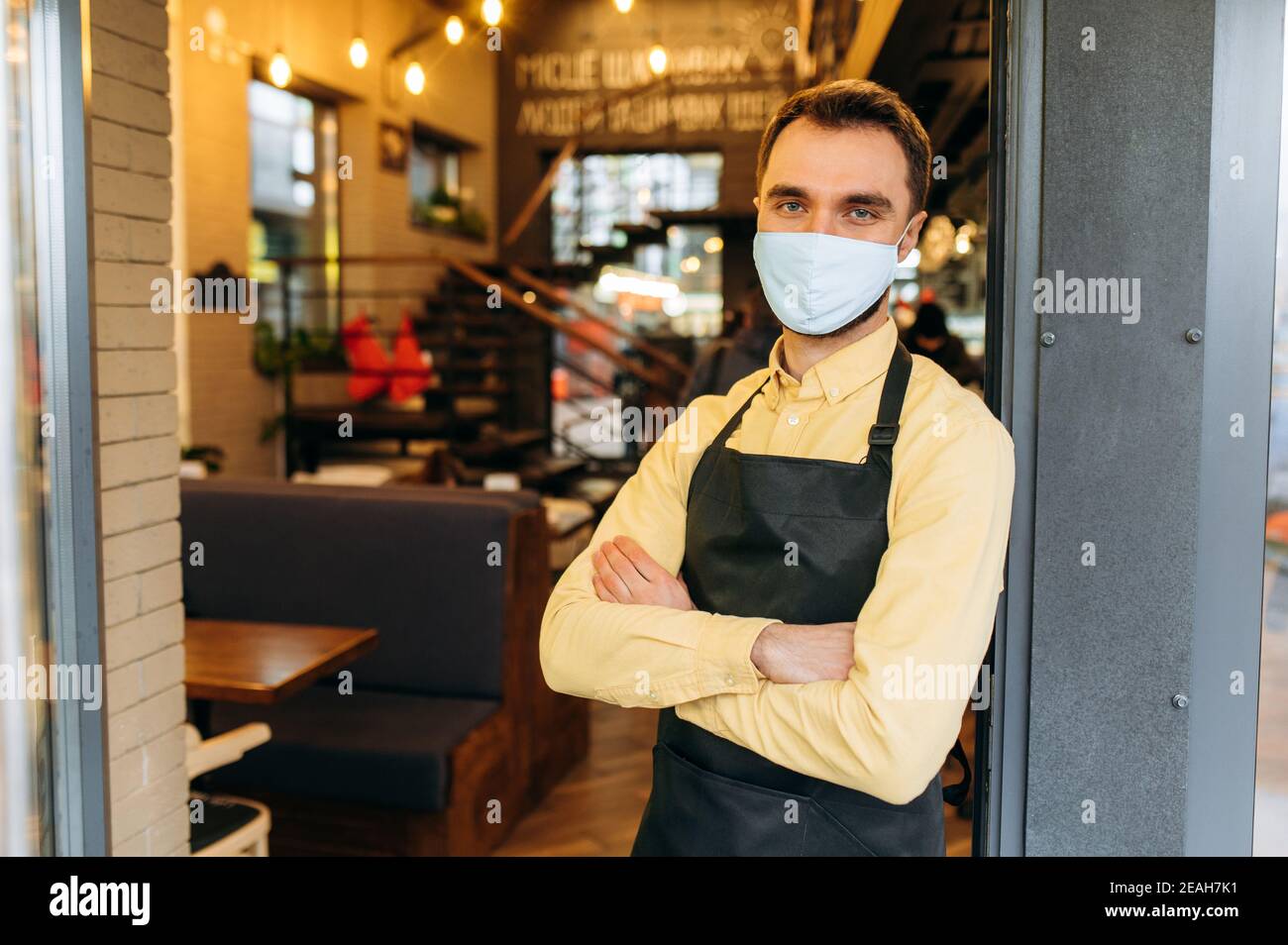 Ein erfolgreicher Kellner, Barista oder Besitzer eines Restaurants, Cafés oder Bars steht am Eingang mit Schutzmaske und schwarzer Arbeitsschürze mit gekreuzten Armen und lädt Gäste ein Stockfoto