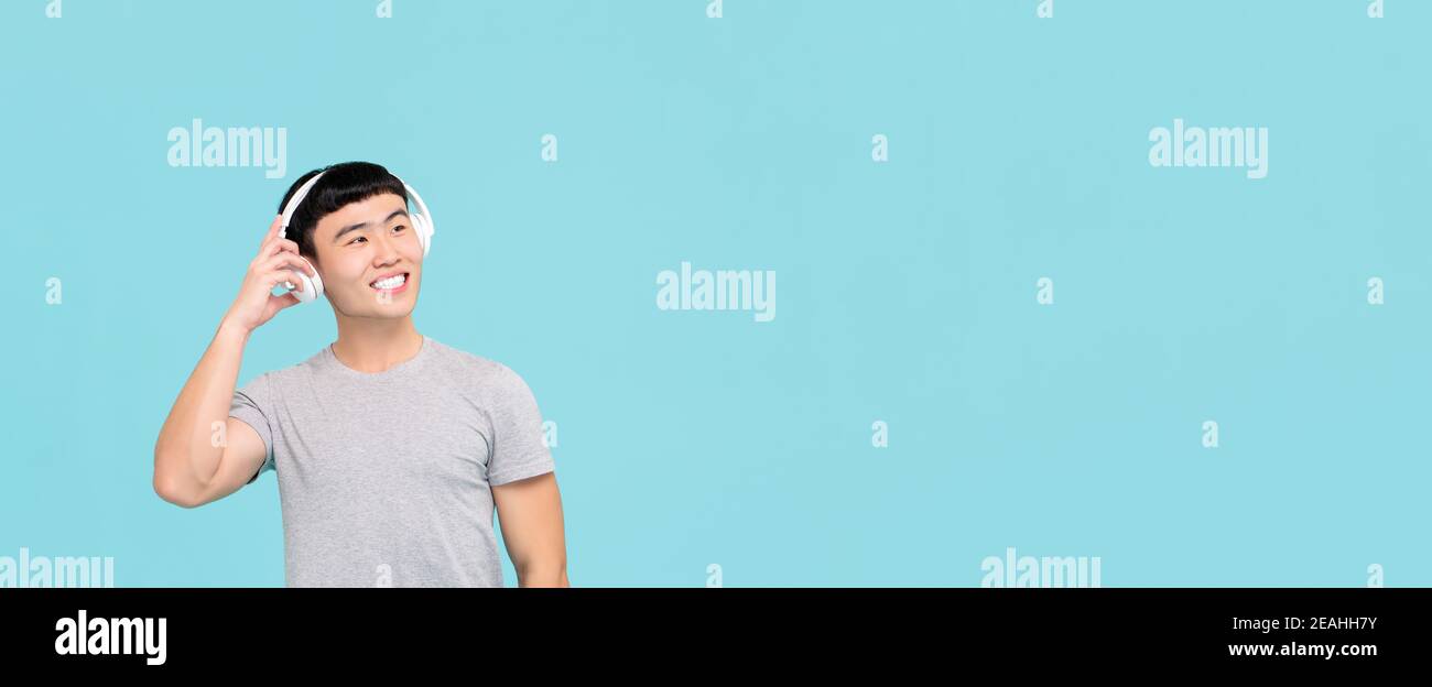Glücklicher junger asiatischer Mann, der kabellose Kopfhörer trägt und Musik hört Und lächelnd auf hellblauem Bannerhintergrund mit Kopierraum Stockfoto