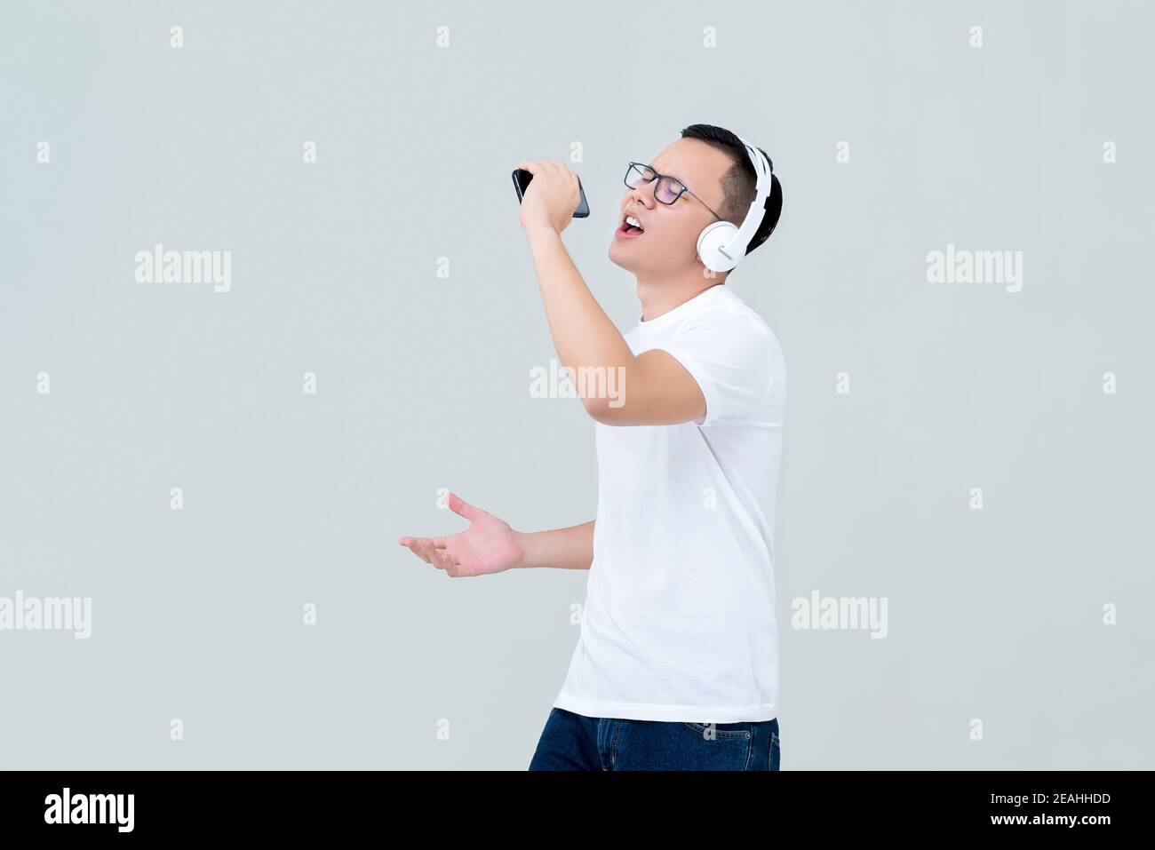Junger asiatischer Mann mit Kopfhörern, der Musik vom Handy hört Telefon und Gesang isoliert auf hellgrauem Hintergrund Stockfoto