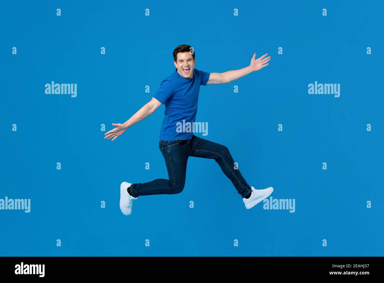 Energischer junger schöner Mann, der mit ausgestreckten Händen springt und lächelt Isoliert auf blauem Hintergrund Stockfoto
