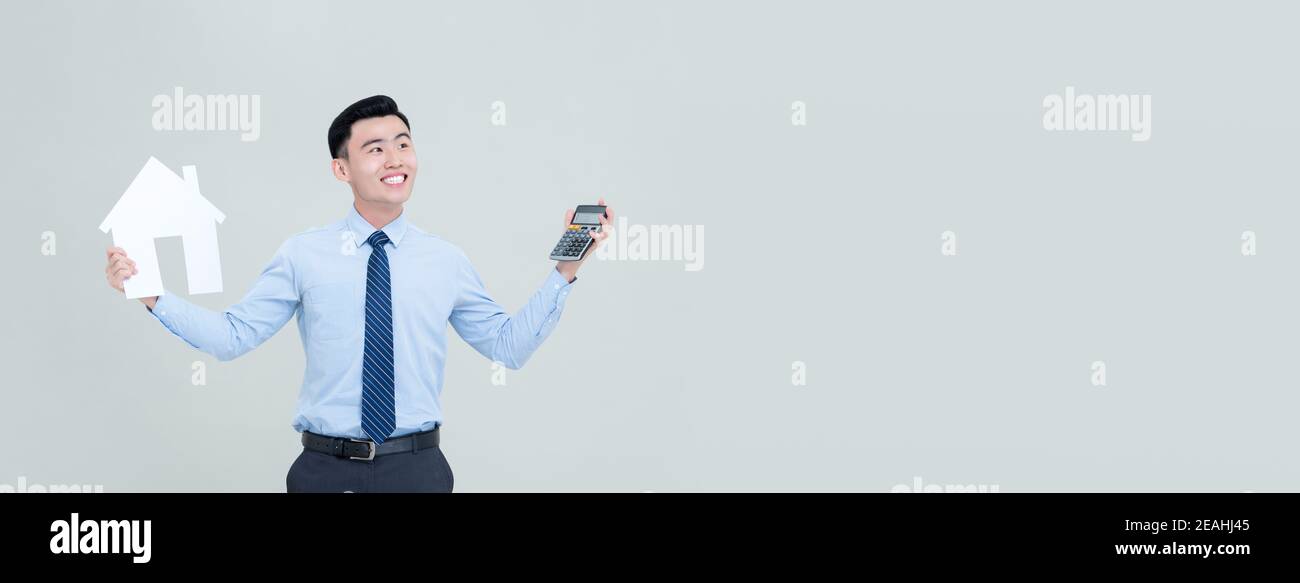Junge lächelnde asiatische männliche Immobilienmakler Holding Rechner und Haus Ausschnitt isoliert auf hellgrauem Banner Hintergrund mit Kopie Platz Stockfoto