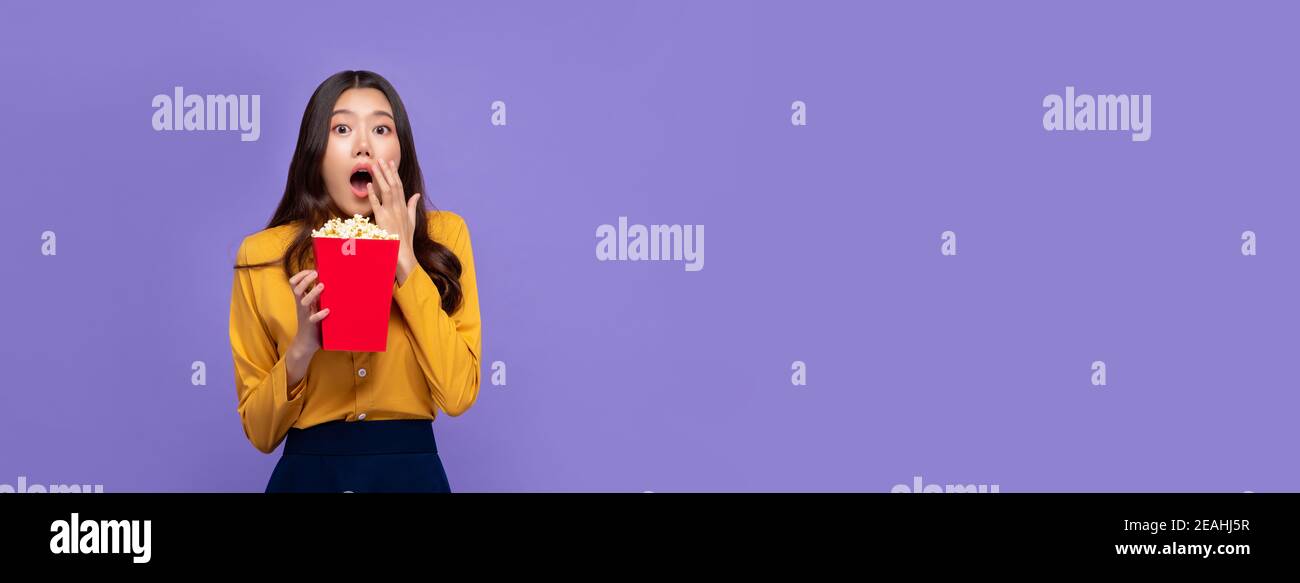 Aufgeregt asiatische Frau hält Popcorn und keuchend mit Handcover Mund beim Ansehen Film isoliert auf lila Banner Hintergrund Stockfoto