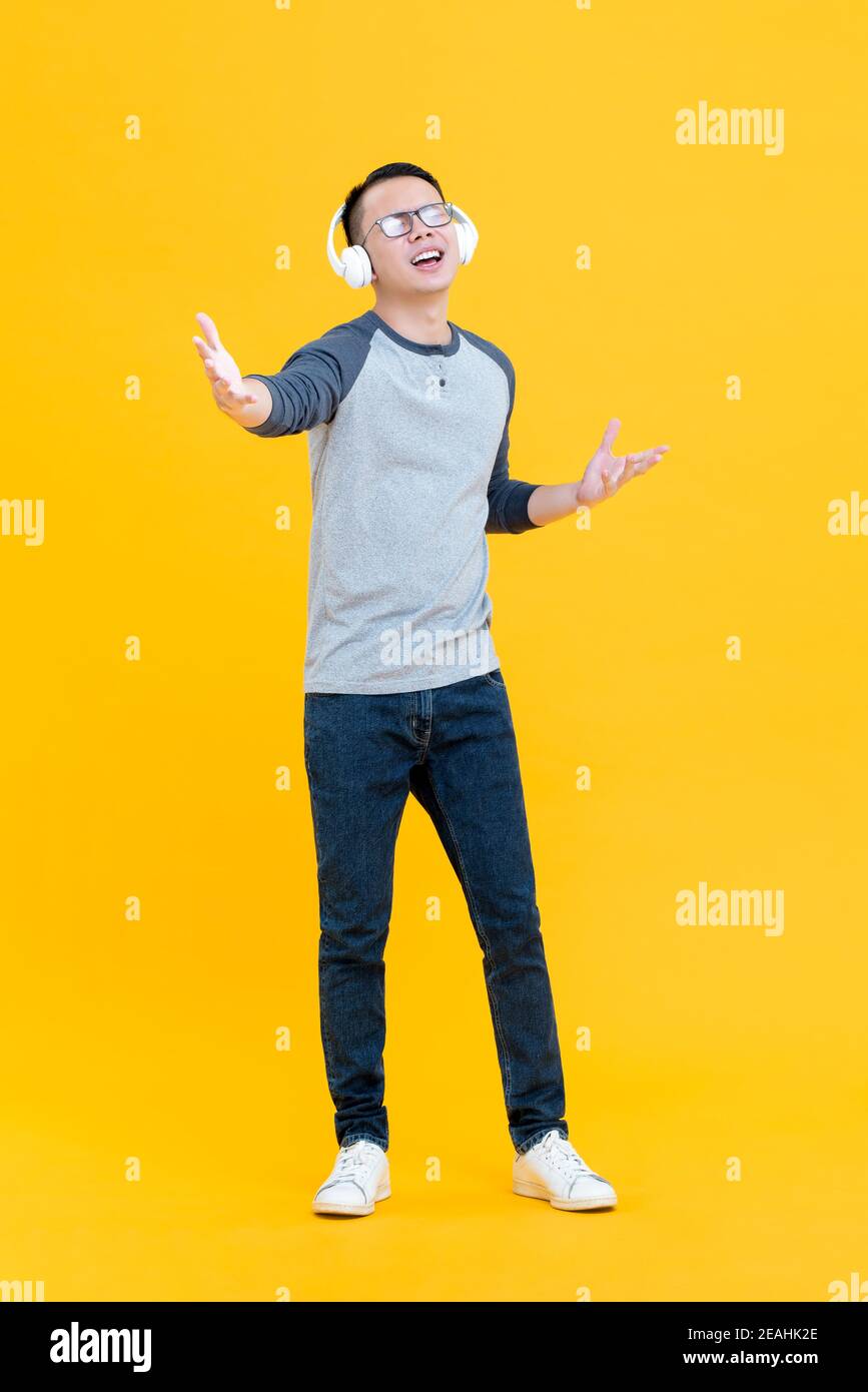 Glücklicher asiatischer Mann genoss das Hören von Musik auf Kopfhörern mit Augen geschlossen und Hände geöffnet isoliert auf gelbem Hintergrund Stockfoto