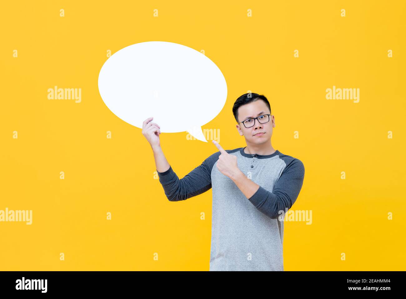 Zweifelhafter asiatischer Mann zeigt auf leere Sprechblase isoliert auf Gelber Hintergrund Stockfoto