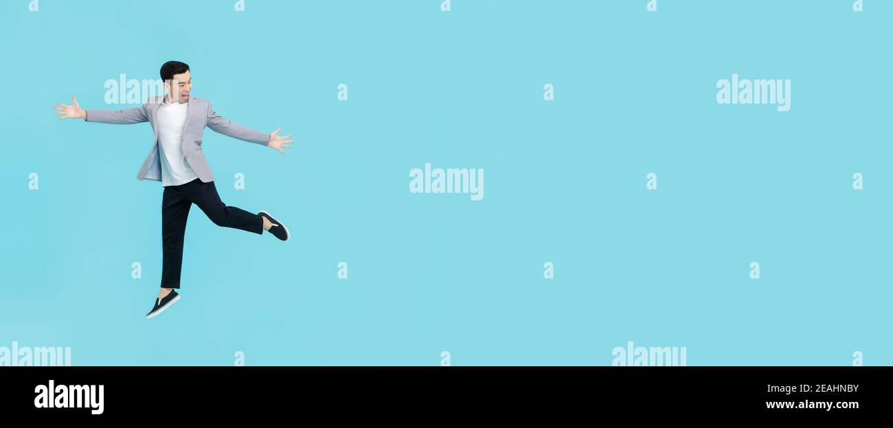 Junge energetische asiatische Mann in smart casual Kleidung outstrecking Hände Und springen mit Begeisterung neben Kopierraum auf hellblau Hintergrund des Banners Stockfoto