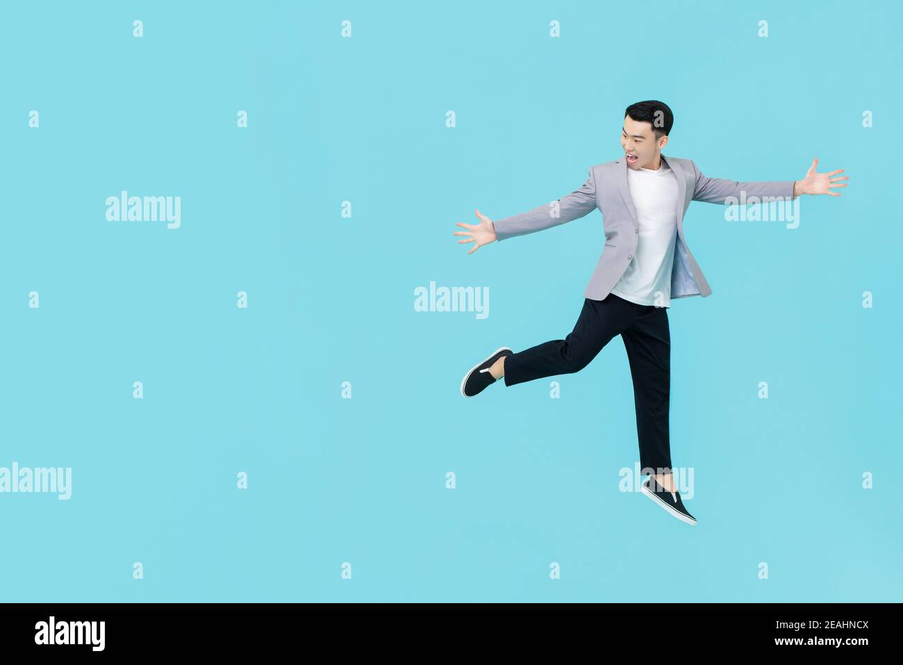 Junge energetische asiatische Mann in smart casual Kleidung outstrecking Hände Und springen mit Begeisterung neben Kopierraum auf hellblau Hintergrund Stockfoto