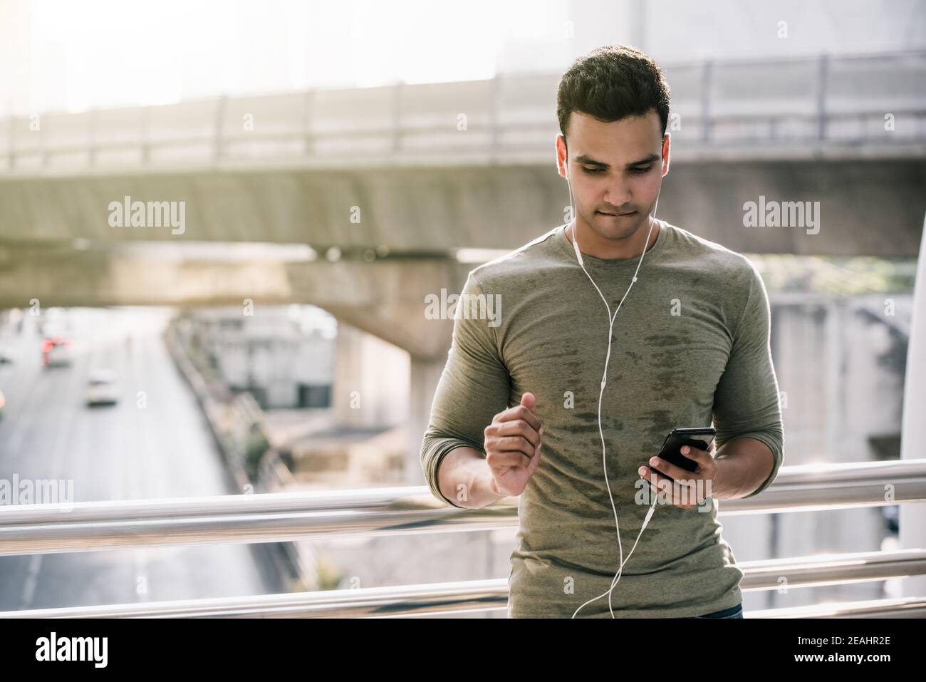 Junge gutaussehende indische Mann in verschwitzten Top Ruhe nach dem Joggen Im Freien und Musik-Streaming auf dem Smartphone gegen Urban hören Verkehrsbehinderungen Stockfoto