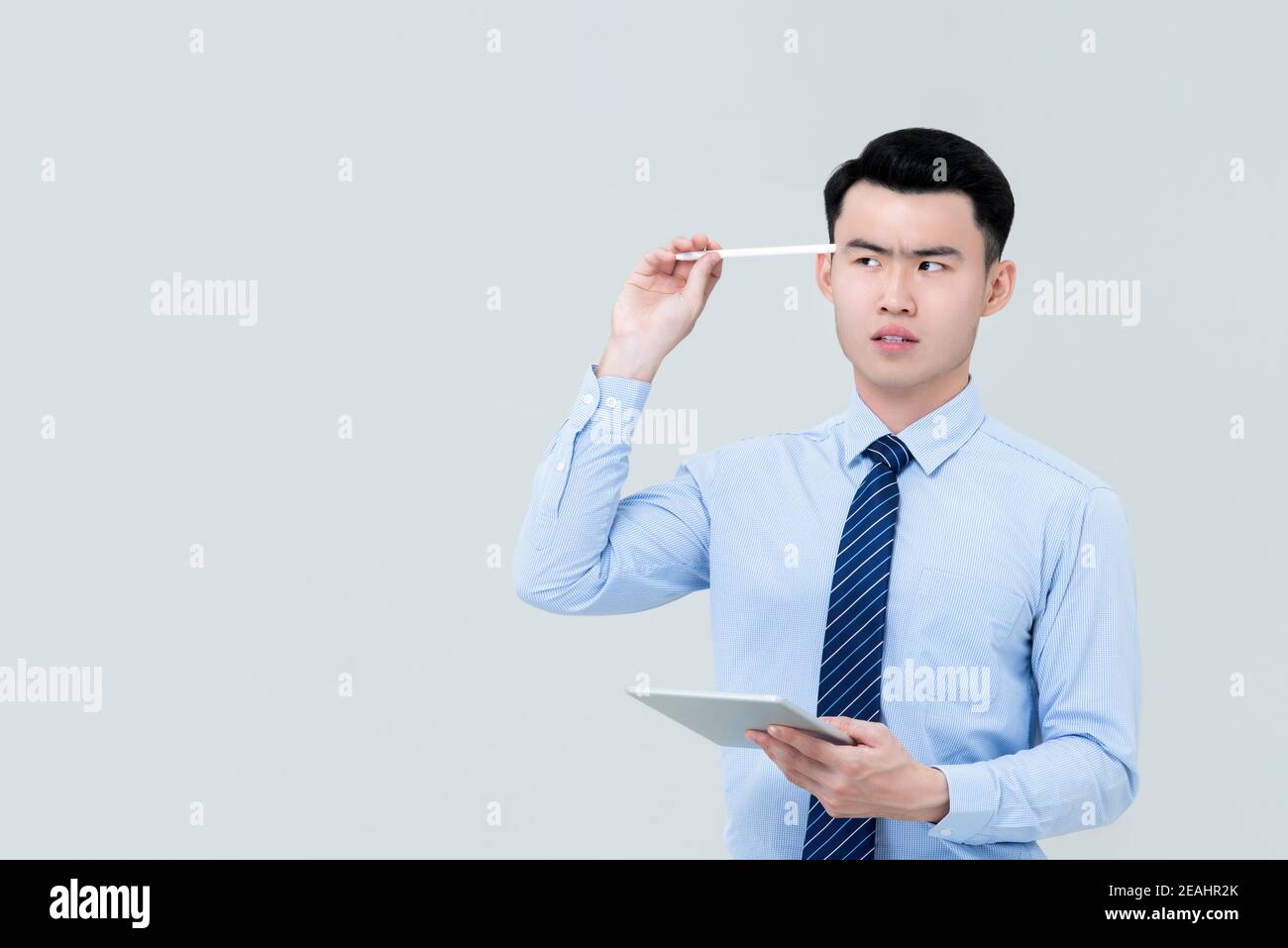 Junge asiatische professionelle Mann in Business-Kleidung suchen intensiv und Durchdachte Blicke seitlich und halten Stift an Kopf mit Tablet In der Hand besid Stockfoto