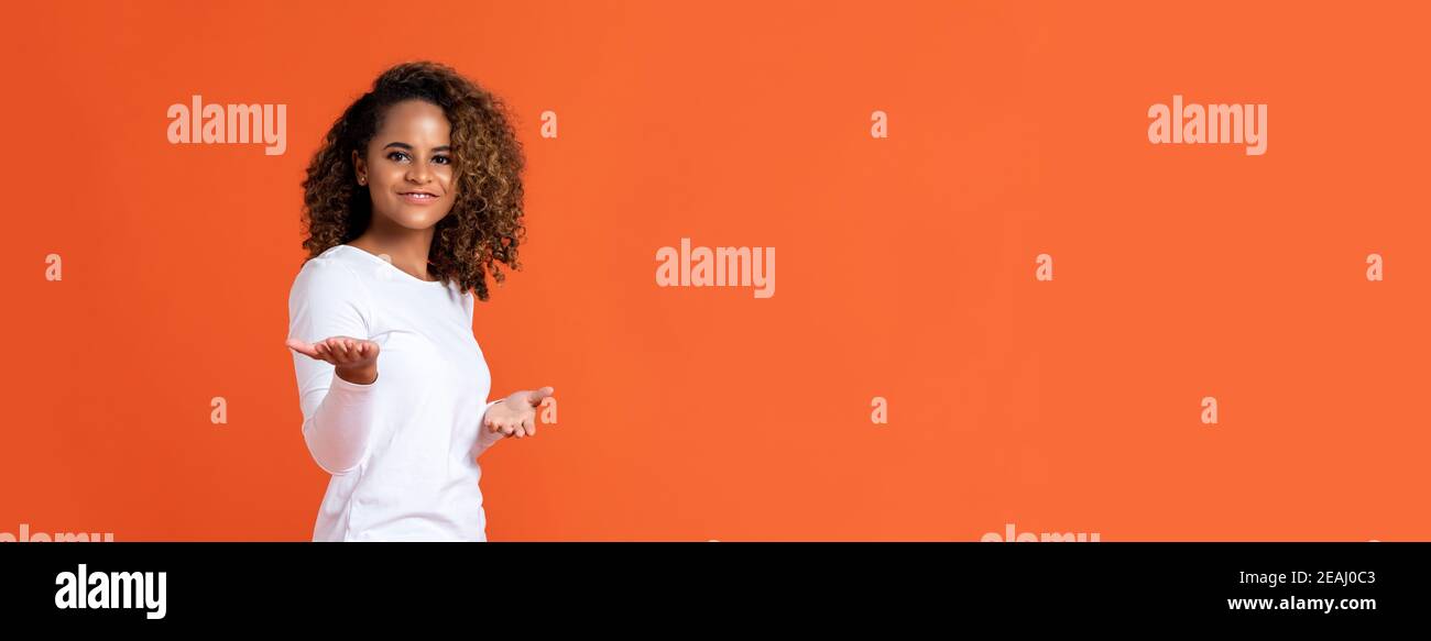 Glücklich lächelnde junge afroamerikanische Frau, die willkommen oder präsentiert Geste mit offenen Händen isoliert auf orangefarbenem Banner Hintergrund mit Speicherplatz kopieren Stockfoto