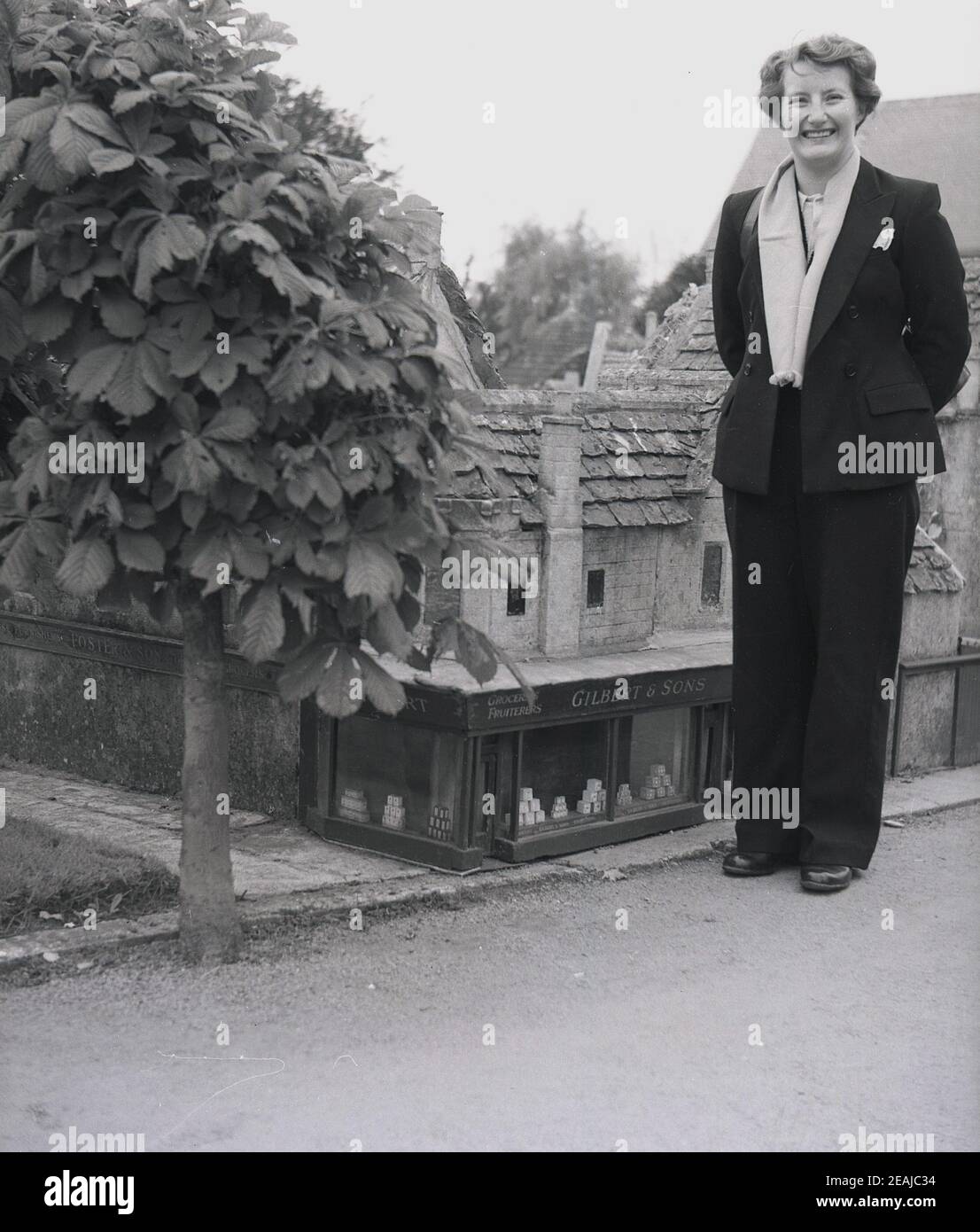 1950s, historisch, eine Dame in einem Modelldorf, das beim Dorflebensmittelgeschäft, Gilbert & Sons, England, Großbritannien, steht. In dieser Zeit hatten viele britische Küstenstädte "Modelldörfer", kleine Kopien von Gebäuden, die ursprünglich als Hobby von einer wohlhabenden lokalen Residenz gebaut und später zu einer Touristenattraktion für Besucher gemacht wurden. Stockfoto