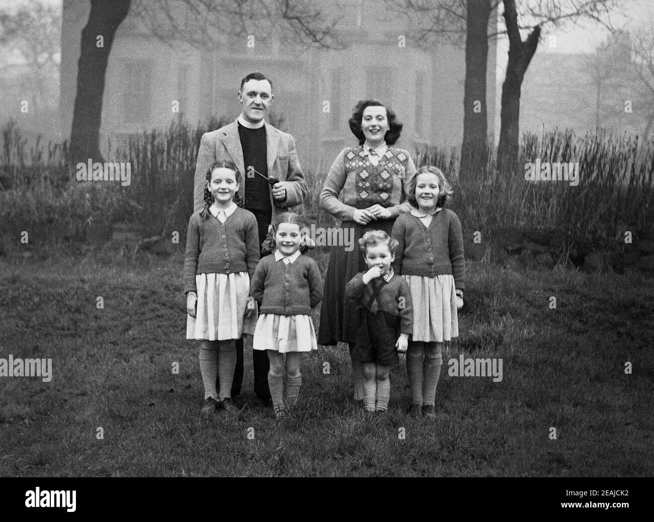 1950s, historisch, ein Familienfoto, draußen auf Gras auf dem Kirchplatz stehend, ein Vikar mit seiner Frau und vier kleinen Kindern mit kleinen Mädchen in der Schuluniform., England, Großbritannien. Der Vikar, der seinen Bürokragenkragen trägt, hält eine lange Pfeife. Stockfoto