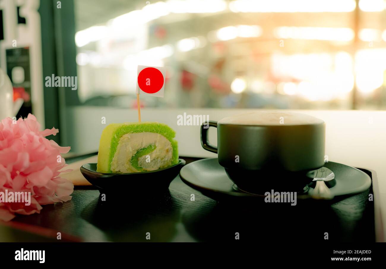 Heiße Kaffeetasse mit Scheibe grüner schweizer Rollkuchen mit Sahne auf dem Teller im Café auf Holzplatte im Café. Latte, Cappuccino oder Espresso im Glas. Heißgetränk. Koffein-Getränk zum Frühstück Stockfoto