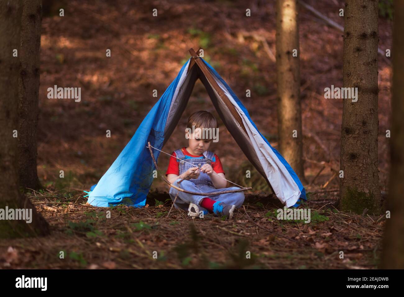Junge sitzt unter einem Spielzelt im Wald, spielt allein, nachdenklich und in Einsamkeit, genießt, einen Bogen zu schaffen, spielen vorgebend. Stockfoto