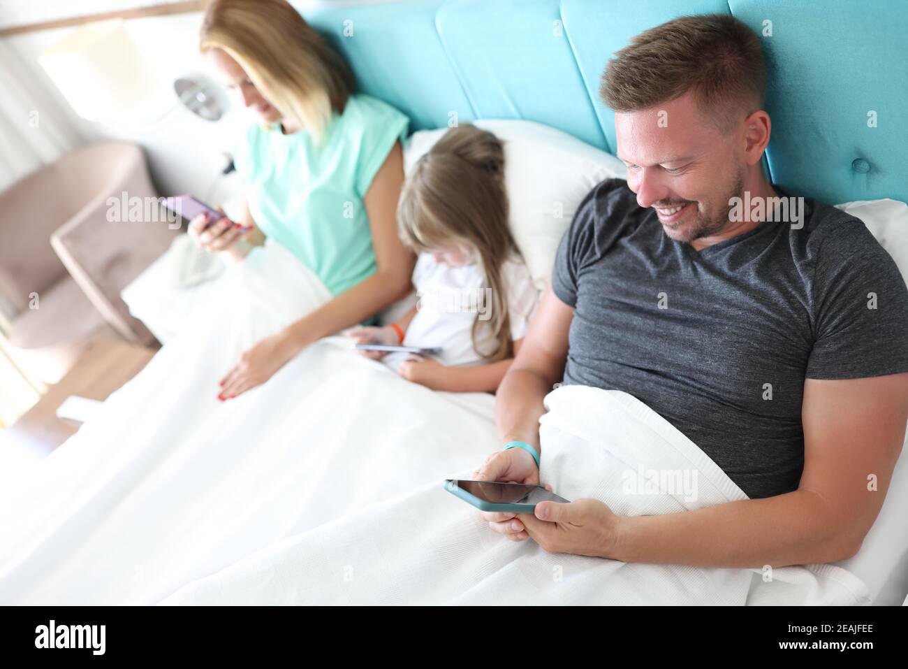 Eltern und Kind liegen im Bett, jeder hält das Smartphone in den Händen Stockfoto