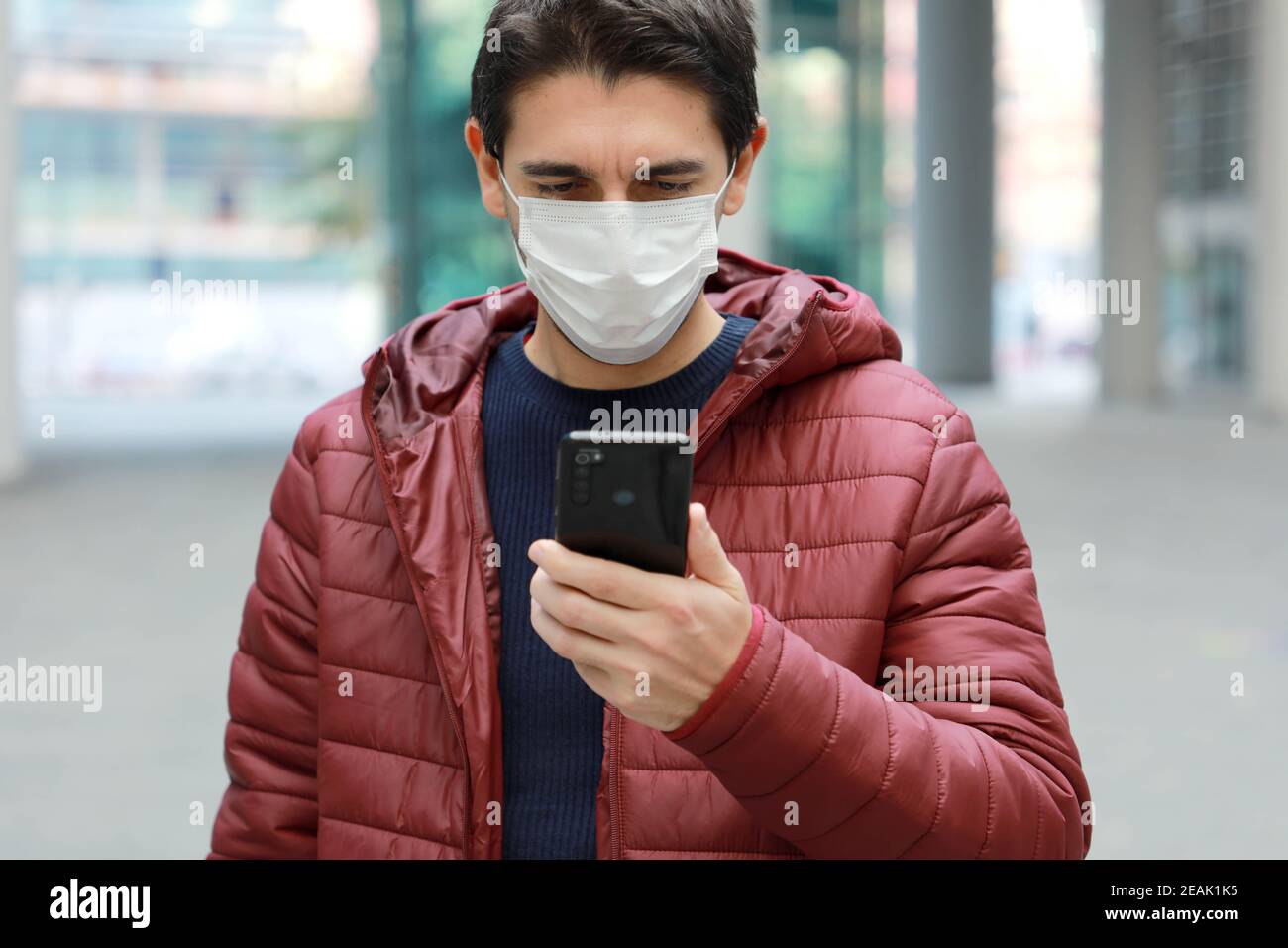 Junger kaukasischer Mann mit Schutzmaske, der Nachrichten oder Nachrichten liest Auf dem Smartphone in der modernen Stadtstraße Stockfoto