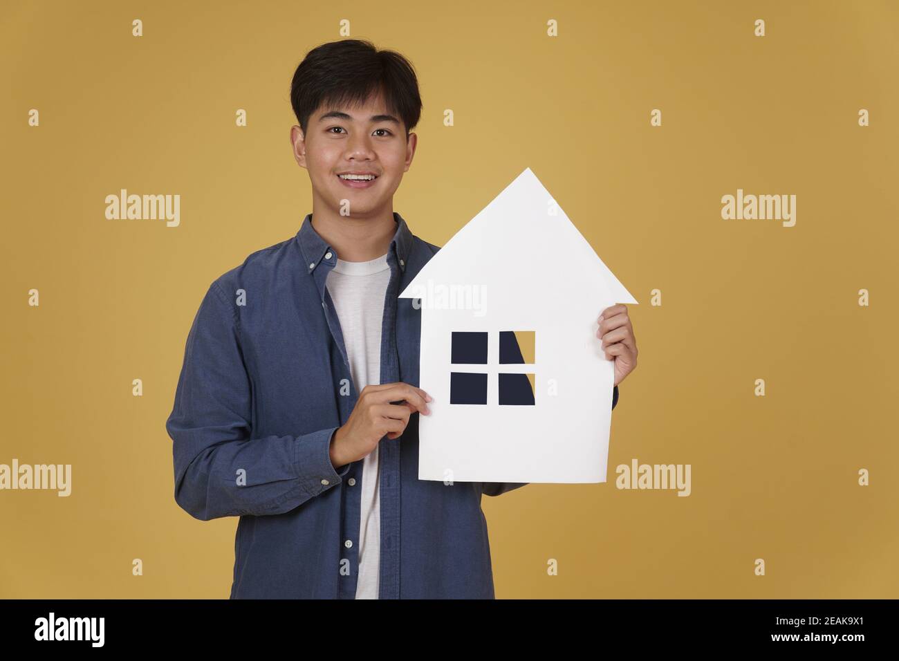 Porträt von lächelnd fröhlich jungen asiatischen Mann lässig mit Haus Papier Ausschnitt isoliert auf gelben Studio-Hintergrund gekleidet. Immobilien pur Stockfoto