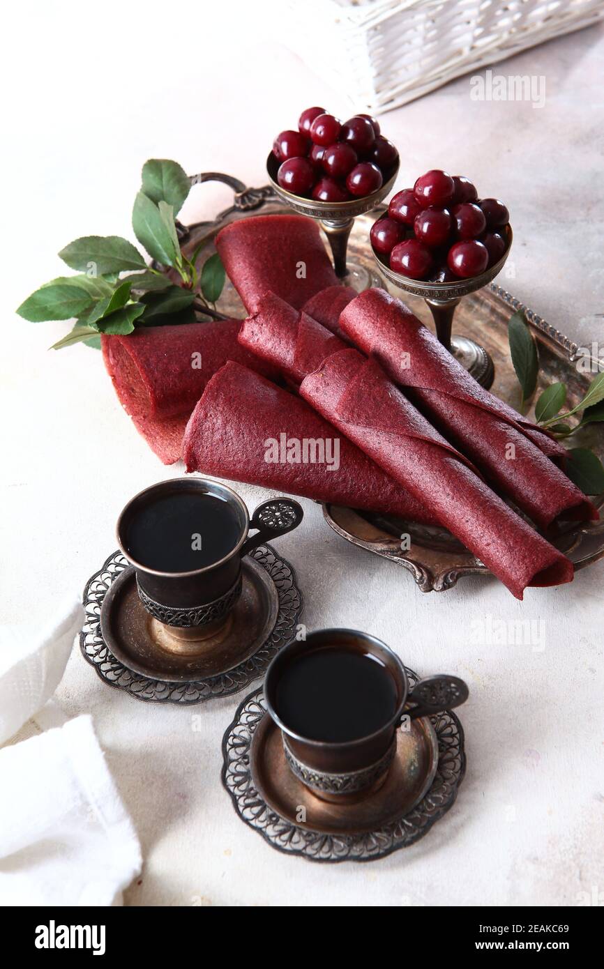 Kirschpastilien werden zu einem Kegel gerollt. Zwei Tassen Kaffee in Vintage-Geschirr. Vertikales Foto. Kirschen und Äpfel sind in einer Schüssel mit Nickel-Silber. Stockfoto