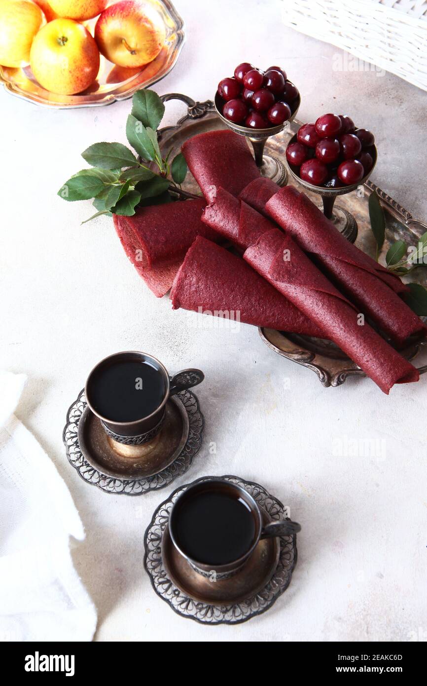 Kirschpastilien werden zu einem Kegel gerollt. Zwei Tassen Kaffee in Vintage-Geschirr. Vertikales Foto. Kirschen und Äpfel sind in einer Schüssel mit Nickel-Silber. Stockfoto