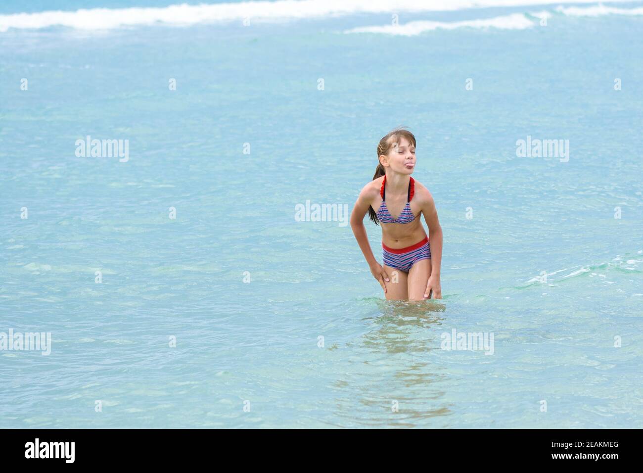 Ein Mädchen steht knietief im Meerwasser, will nicht zum Sonnen ausgehen und streckt ihre Zunge aus Stockfoto
