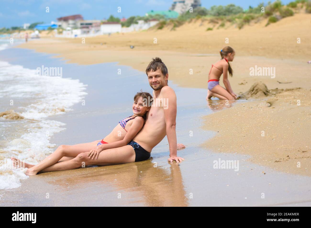 Papa und Tochter sitzen im Vordergrund, im Hintergrund baut ein Mädchen eine Sandburg Stockfoto