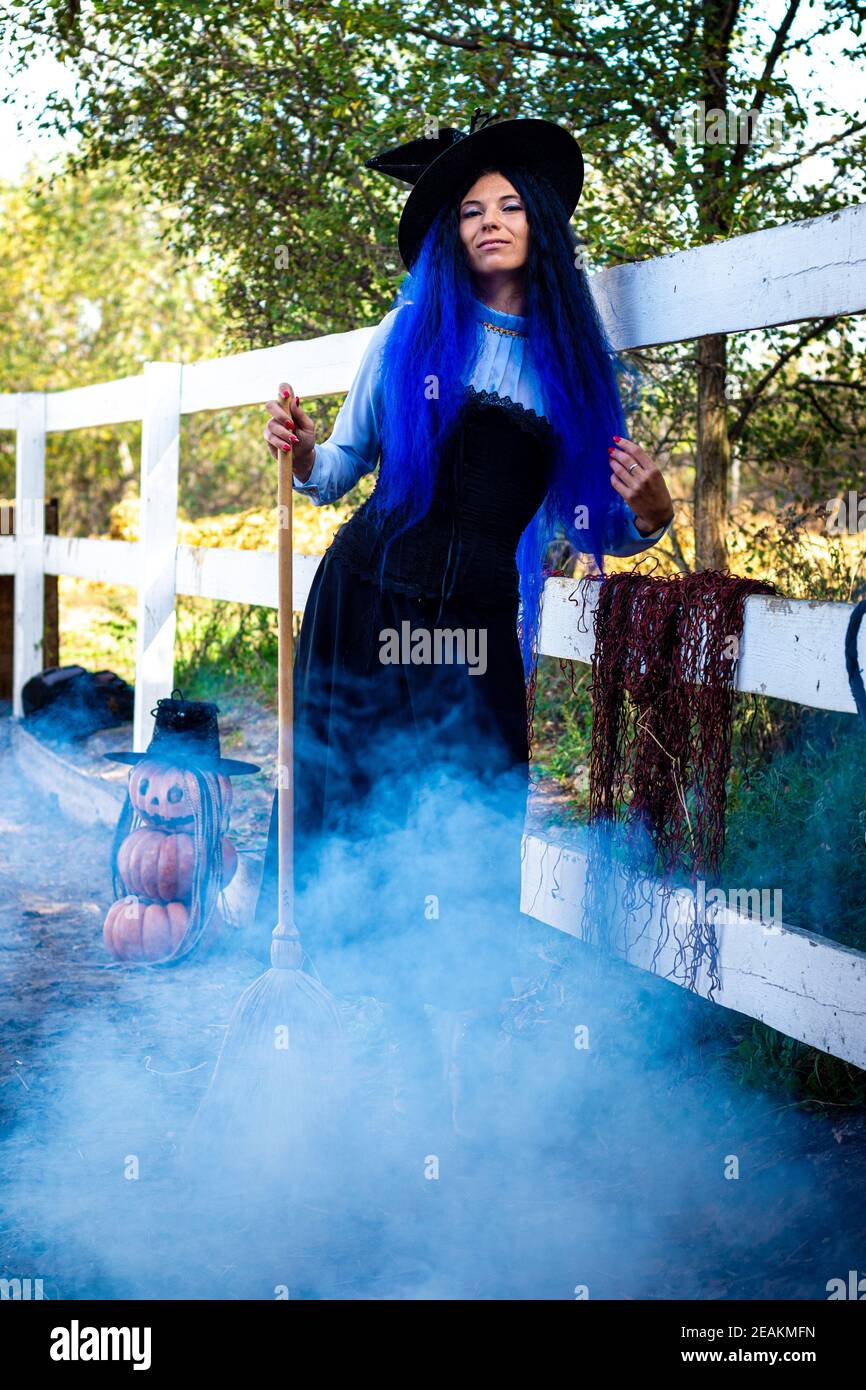 Ein Mädchen als Hexe gekleidet steht an einem Zaun auf einer Farm, Rauch von unten, Kürbisse im Hintergrund Stockfoto