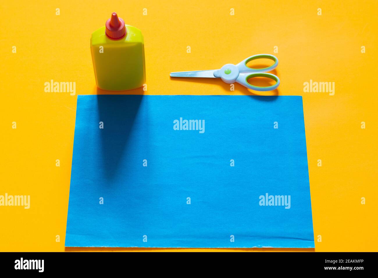 Auf einem gelben Hintergrund liegt ein Blatt blaues Papier, daneben befindet sich eine Tube Leim und eine Schere Stockfoto