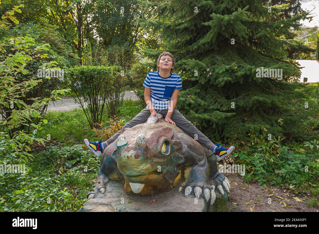 Fröhlich lächelnder Junge in einem gestreiften T-Shirt sitzt auf einem Dinosaurierstatue im Park auf dem Spielplatz Stockfoto