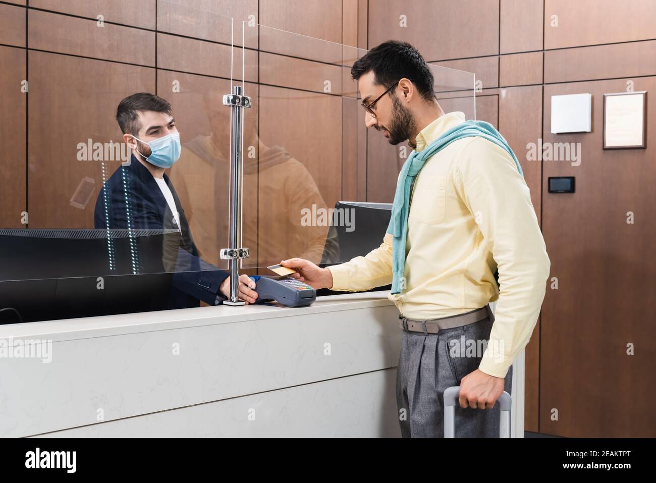Rezeptionist in medizinischer Maske hält Zahlungsterminal in der Nähe muslimischer Mann Mit Koffer und Kreditkarte in der Hotellobby Stockfoto