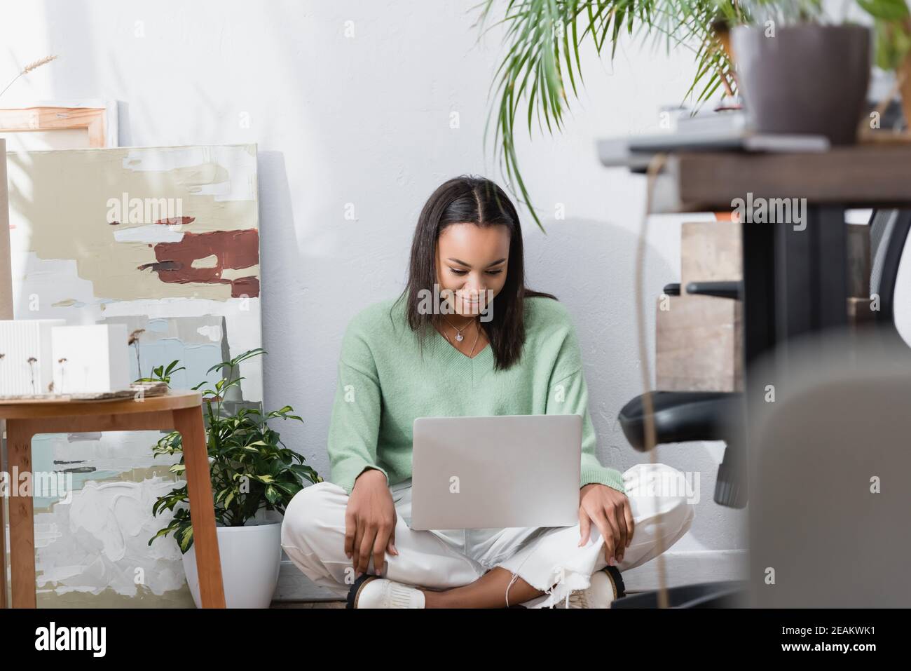 Glücklicher afroamerikanischer Architekt, der lächelt, während er auf dem Boden sitzt Laptop im Heimstudio auf verschwommenem Vordergrund Stockfoto