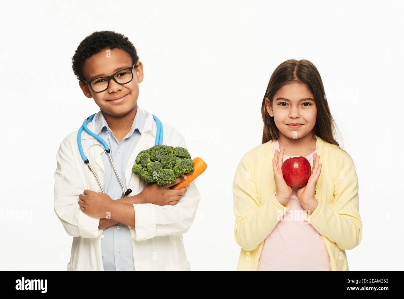Junge in Ärzteuniform stehend mit einem Mädchen, halten sie gesündeste Gemüse. Benefits Brokkoli, Apfel, Karotte für Kinder Stockfoto