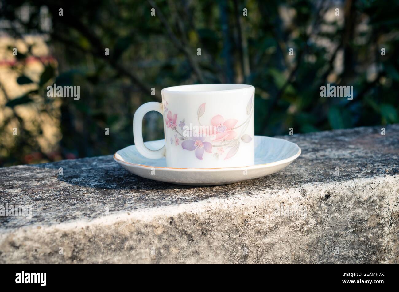 Kaffeetasse im Morgenlicht. Sommerlich frischer cooler Look. Weiße Kaffeetasse auf Untertasse für Heißgetränk auf Dachbalken eines Wohnhauses mit Bokeh Hintergrund. Stockfoto