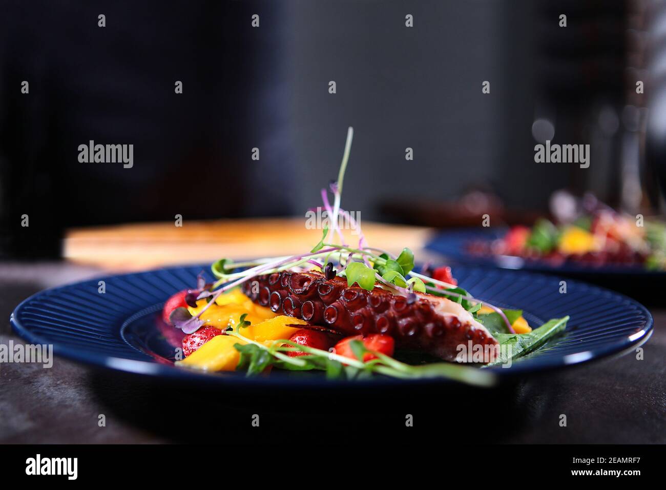 Octopus Salat mit Mango und Erdbeeren auf einem blauen Teller. Microgreens in einem Salat mit einem Oktopus. Nahaufnahme. Stockfoto