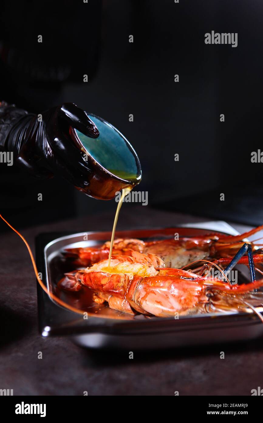 Der Koch gießt Sauce auf die gegrillten Garnelen. Riesige Süßwassergarnelen. Gegrillte Garnelen. Vertikales Foto auf schwarzem Hintergrund. Nicht erkennbare Person. Kochkonzept. Stockfoto