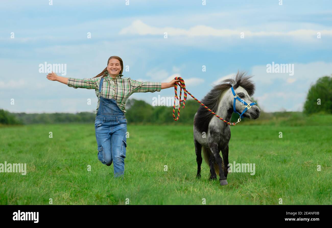 Lächelnde kaukasische Frau läuft mit weit ausgestreckten Armen. Erwachsene weibliche Trainerin im karierten Hemd und blauen Jeans und ihr Pony sind in ländlichen. Stockfoto