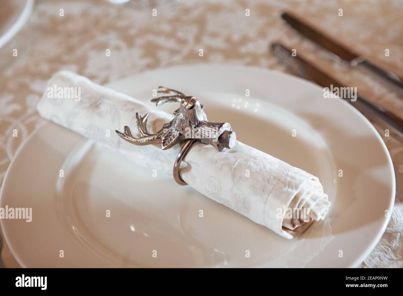 Die weiße Stoffserviette wird mit einem Ring in Form eines Hirschkopfes gesichert. Gabel und Messer aus dem Fokus.der Blick von oben. Makrofoto. Speicherplatz kopieren. Stockfoto