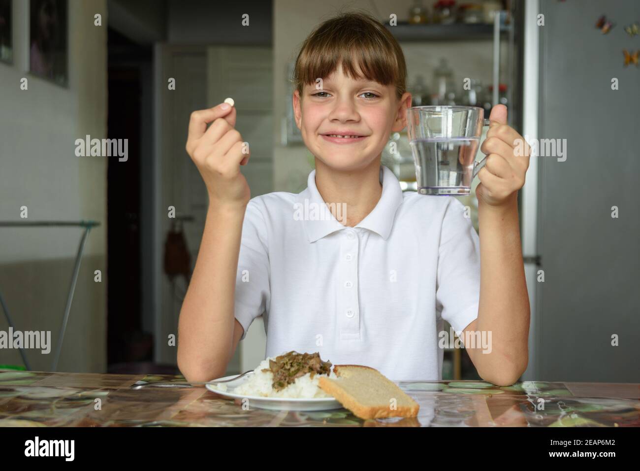 Glückliches Mädchen am Esstisch hält eine Pille in der einen Hand, ein Glas Wasser in der anderen Stockfoto
