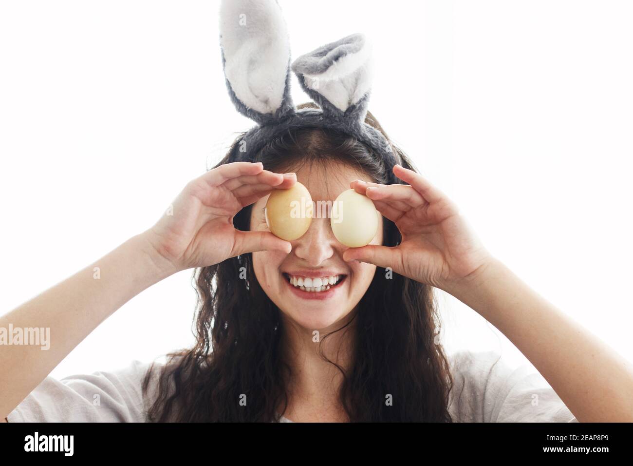 Frohe Ostern! Glückliche junge Frau in Hasenohren und Leinenkleid hält ostereier an den Augen und lächelt in weißem Licht. Natürlich gefärbte ostereier in der Hand Stockfoto