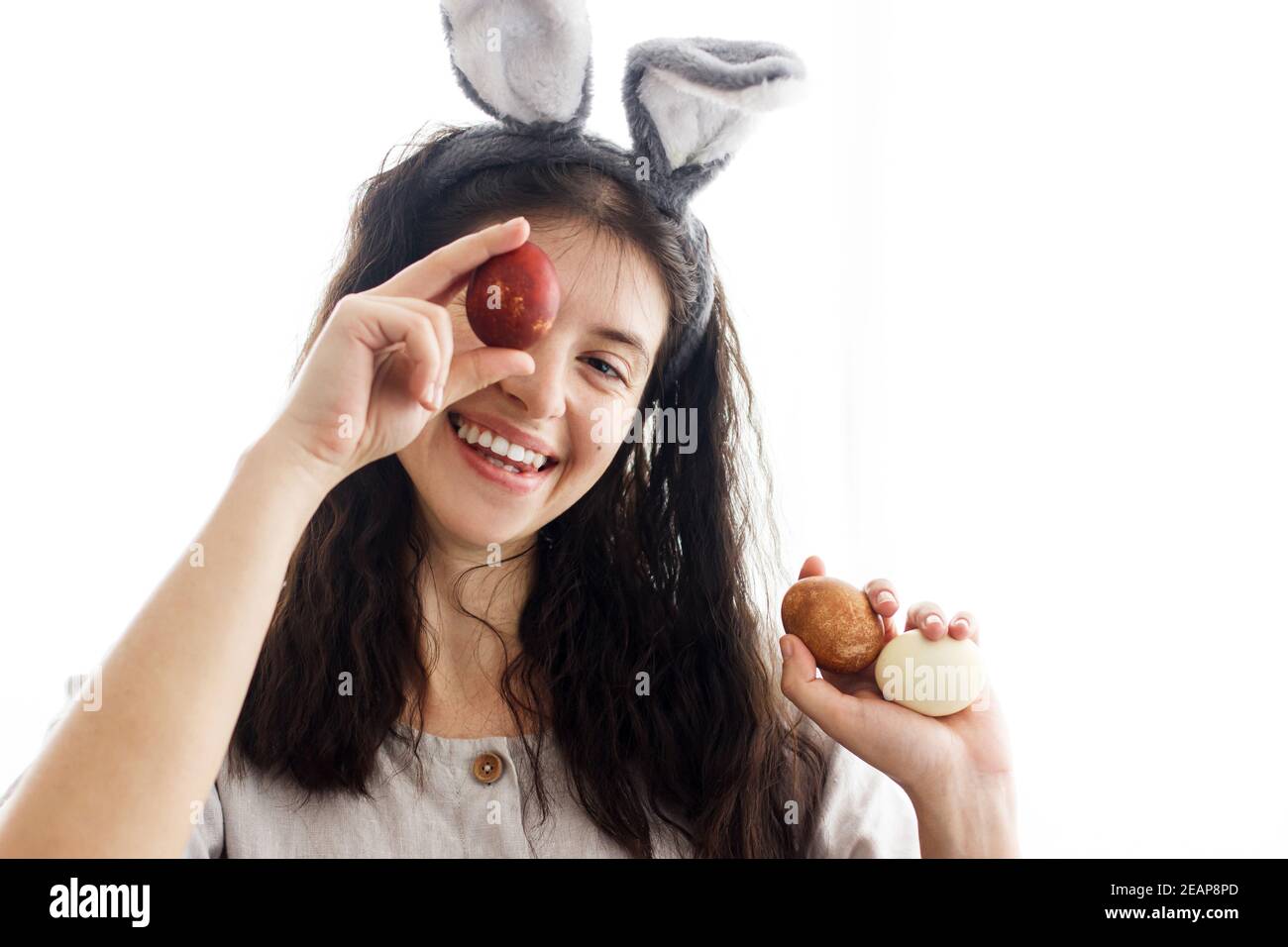 Frohe Ostern! Glückliche junge Frau in Hasenohren und Leinenkleid hält ostereier an den Augen und lächelt in weißem Licht. Natürlich gefärbte ostereier in der Hand Stockfoto