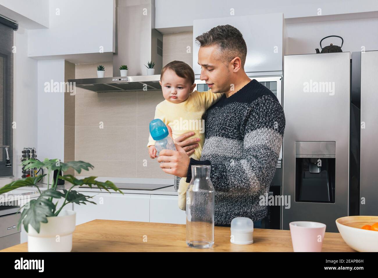 Stock Foto von Young Single Dad Vorbereitung einer Flasche Fütterung, während er sein Baby in den Armen zu Hause. Mann und Sohn, Elternschaft Vaterschaft Moment. Einzelne da Stockfoto