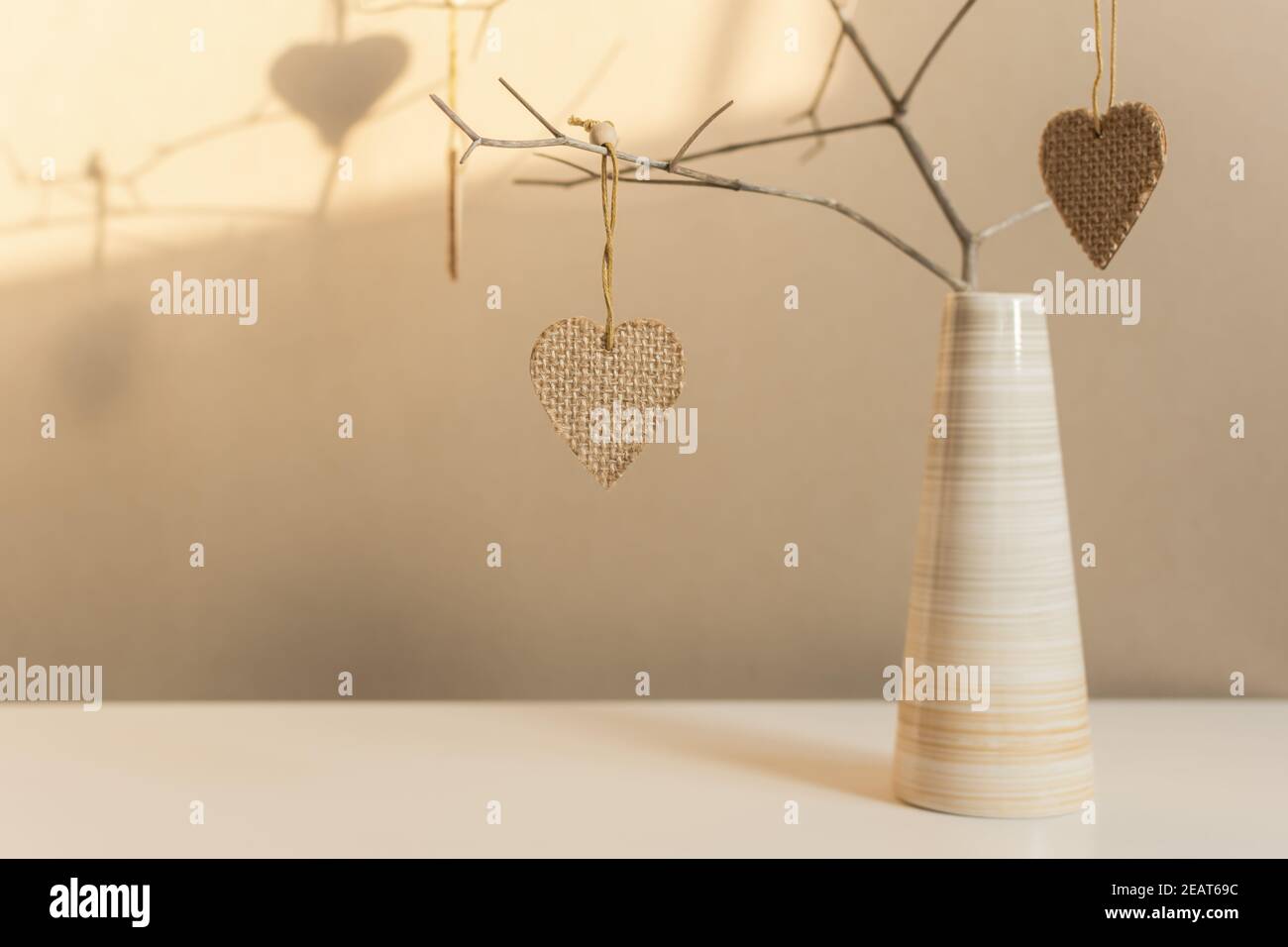 Kreative Komposition in neutralen Farben zum Valentinstag. Herzen auf einem Zweig in einer Vase auf dem Tisch. Handgefertigte, grobe Quaderlappen-Qualität. Warme Atmosphäre Stockfoto