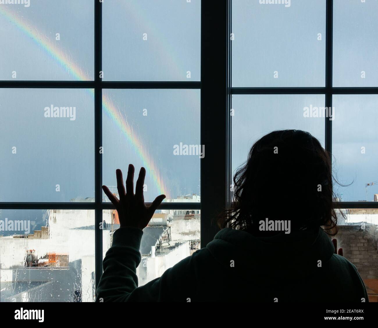 Frau, die am regnerischen Tag aus dem Fenster in Richtung Regenbogen schaut. Conept Bild; weibliche Depression, häuslicher Missbrauch, Selbstisolation, Quarantäne, Coronavirus, Stockfoto
