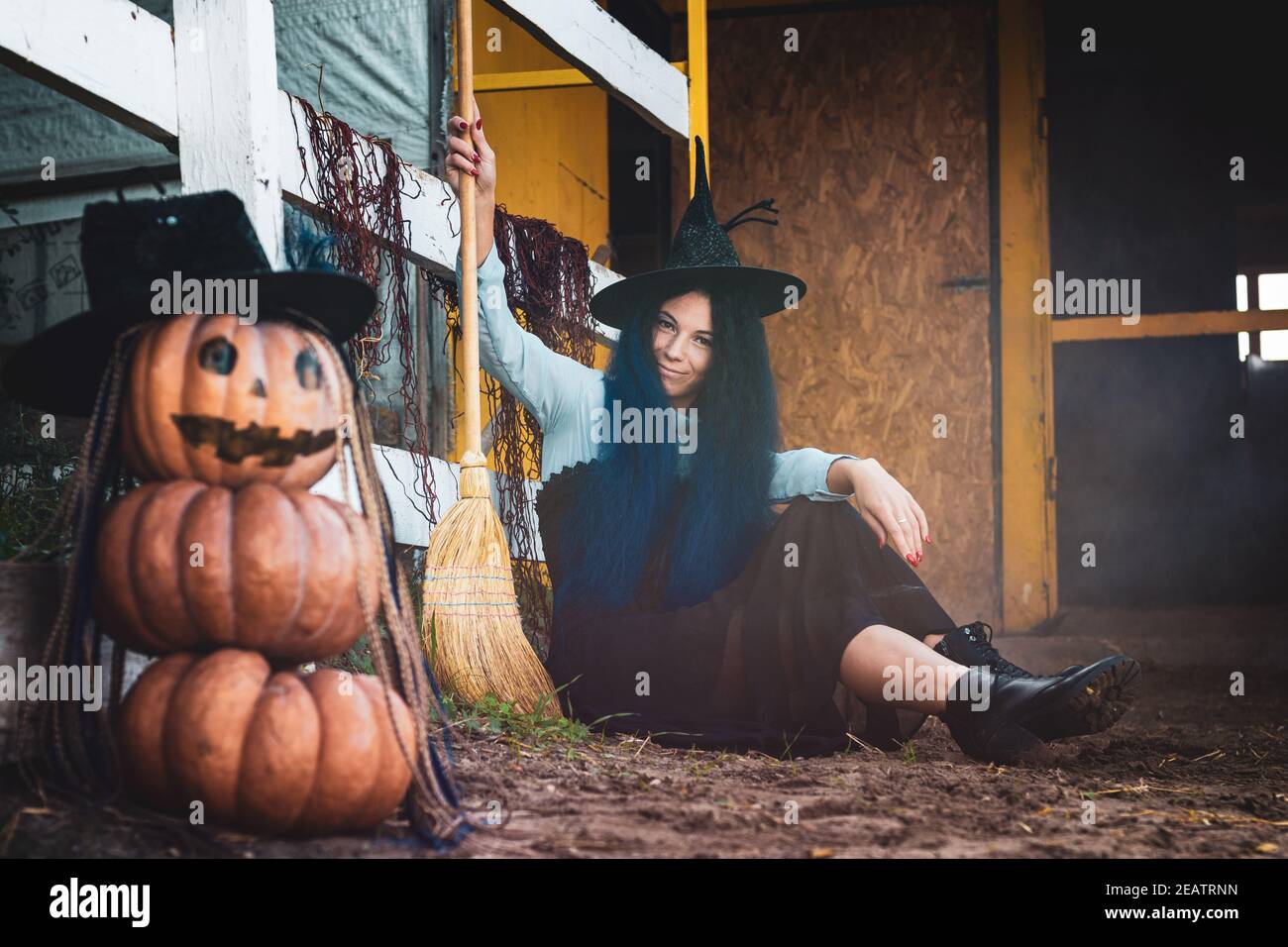 Ein Mädchen als Hexe gekleidet sitzt an einem Zaun mit einem Besen in den Händen, im Vordergrund eine gruselige Figur von Kürbissen Stockfoto