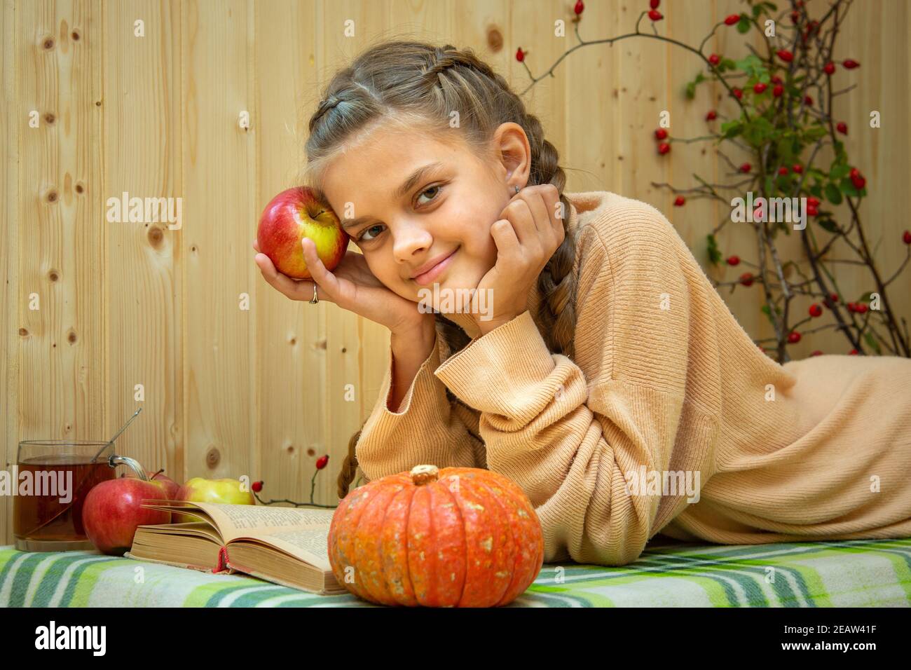 Ein Mädchen, das sich hinlegt, liest ein Buch, ein Kürbis und Äpfel liegen in der Nähe, ein Mädchen schaut freudig in den Rahmen Stockfoto