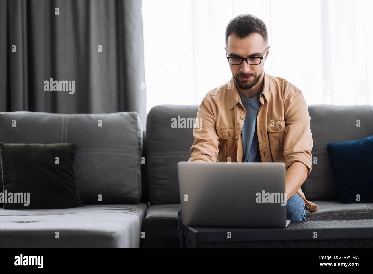 Konzentrierter männlicher Freiberufler, der von zu Hause aus arbeitet und Informationen im Netzwerk sucht. Kaukasische männliche Manager oder IT-Spezialist sitzen auf der Couch, uns Stockfoto