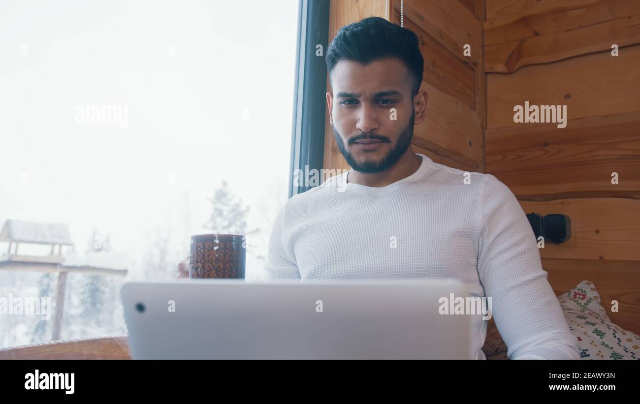 Junger inder trinkt Kaffee und arbeitet am Laptop in der Nähe des großen Fensters. Hochwertige Fotos Stockfoto