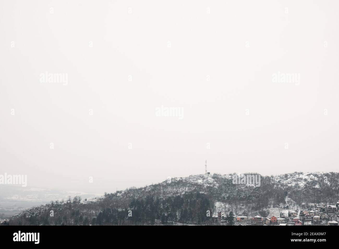 Minimalistische Aussicht mit viel negativem Raum von Schnee bedeckt felsigen Hügel mit Bäumen und TV-Antenne an der Spitze, steigt über kleine Gruppe von Häusern Stockfoto